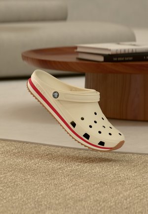 Beige Crocs sko med rød stribe, ventilationshuller og justerbar hælstrop, svævende over et tæppet gulv nær et træbord.