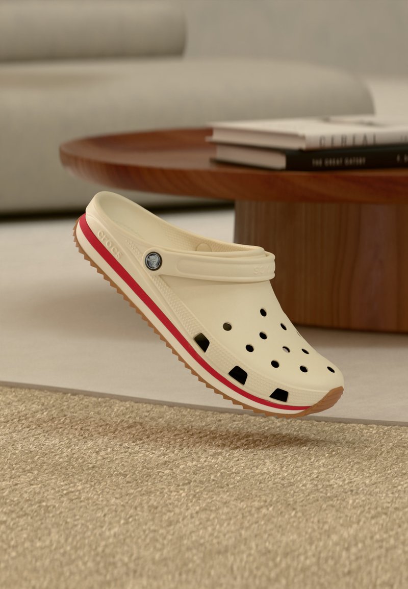 Chaussure Crocs beige avec une bande rouge, des trous de ventilation et une bride de talon réglable, flottant au-dessus d’un sol moquetté près d’une table basse en bois.