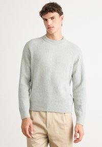 Pull en maille côtelée gris clair avec un col rond, des manches raglan longues et des fentes sur les côtés. Sa texture douce s'associe à une coupe décontractée.