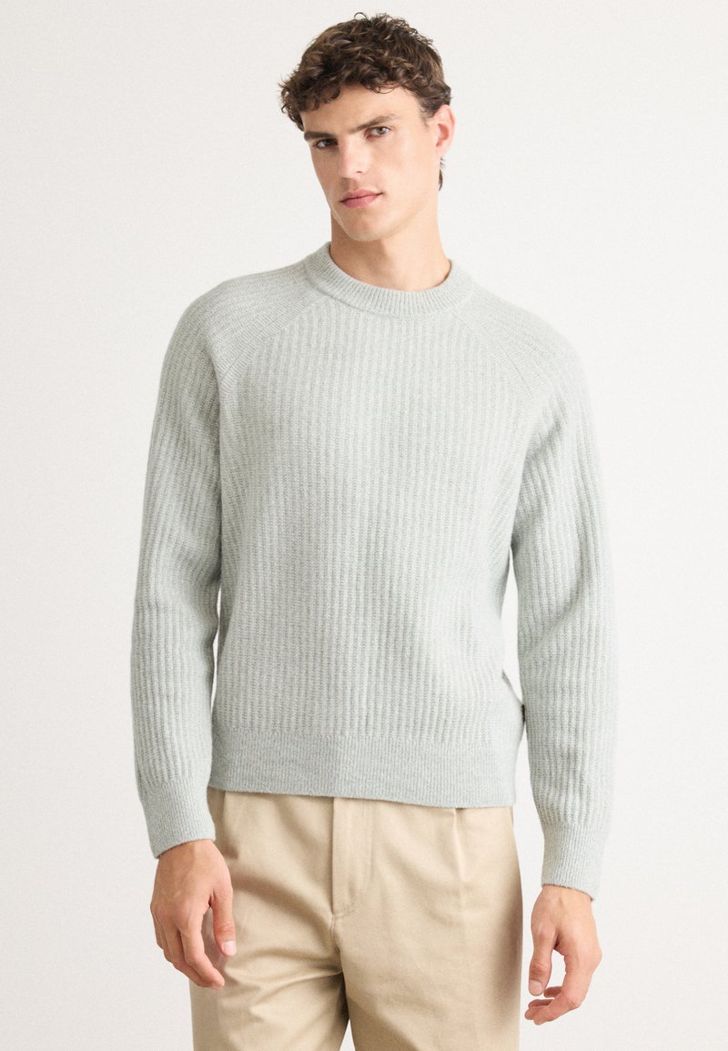 Pull en maille côtelée gris clair avec un col rond, des manches raglan longues et des fentes sur les côtés. Sa texture douce s'associe à une coupe décontractée.