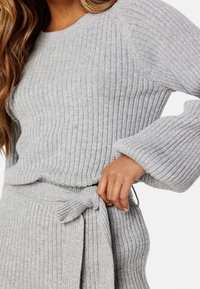 Haut en tricot gris à côtes avec des manches longues évasées et un détail de taille nouée. Le tissu a une finition douce et texturée avec des lignes horizontales.