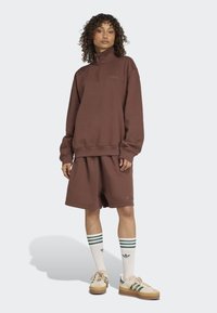 Žena s kučeravými vlasmi, oblečená v hnedém mikinovém svetriku s čtvrtinovým zipem od Adidas, zodpovedajících šortkách, bielych ponožkách Adidas s zelenými pruhmi a béžových teniskách.