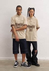 Beige polo skjorter med brune detaljer, med grafiske design. Ett par i mørke denimshorts, det andre i svarte slitne bukser og støvler.