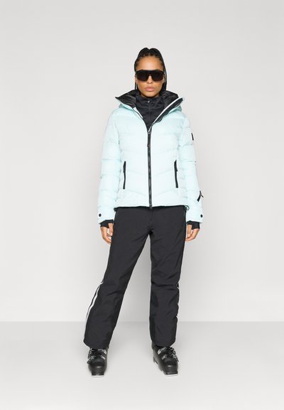 Ανοιχτό μπλε μπουφάν τύπου puffer με μαύρες λεπτομέρειες, φερμουάρ και κουκούλα. Συνδυασμένο με μαύρα σκι παντελόνια με λευκές ρίγες και μαύρες μπότες σκι.