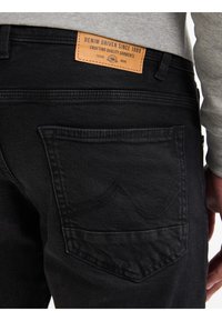 Primer plano del bolsillo trasero de unos vaqueros de denim negro con detalle de costura y etiqueta de cuero marrón que dice "Denim impulsado desde 1989."