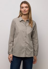 Koszula na guziki z jasnobrązowego materiału z cienkimi, pionowymi białymi paskami; posiada kołnierzyk i długie rękawy, zestawiona z niebieskimi dżinsami.