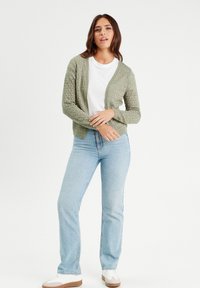 Hellgrüner gestrickter Cardigan mit cropped Design, Rippkante und strukturiertem Muster, kombiniert mit hellblauen Jeans und weißen Sneakers.