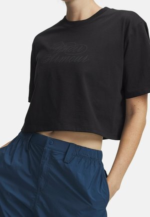 Person trägt ein schwarzes, kurz geschnittenes T-Shirt mit schwachem Text und hoch taillierte, lockere blaue Hose mit elastischem Bund und Seitentaschen.