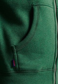 Felpa verde con un tessuto morbido e texturizzato. Presenta una tasca frontale e una cerniera. Maniche e fondo a costine, con un piccolo etichetta del logo visibile.