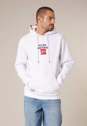 Homme portant un sweat à capuche blanc avec l'inscription "It's the real thing" et le logo Coca-Cola, les mains dans les poches, regardant vers la droite sur un fond beige uni.