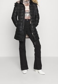 Černý zateplený kabát se zipem a kapucí, s lesklým povrchem, dlouhými rukávy a bočními kapsami, spárovaný s rozšířenými černými kalhotami a bílými botami.