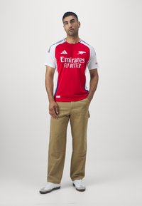 Maillot rouge et blanc à manches courtes avec le logo "Emirates Fly Better", associé à un pantalon cargo beige et des baskets blanches.
