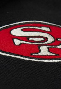 Schwarzer Stoff mit einem auffälligen roten und weißen gestickten Logo, das die Buchstaben "S" und "R" zeigt, umrandet mit schwarzem Stich.