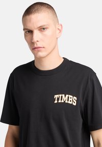 Sort bomulds-t-shirt med korte ærmer og rund hals, med "TIMBS" i beige bogstaver på venstre bryst. Glat tekstur.
