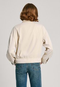 Veste courte de couleur crème avec un tissu lisse et des épaules tombantes ; associée à un jean bleu avec un design classique à 5 poches.