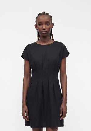 Femme aux cheveux tressés portant une robe noire simple à manches courtes, cintrée à la taille, debout devant un fond blanc uni.