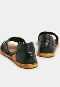 Naha musta nahka sandaalit, joissa on kudottu päällysosa, avoin takapää ja vaaleanruskea pohja, jossa on ompeluyksityiskohtia. Tasapohjaiset ja helposti jalkaan laitettavat.