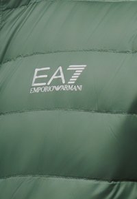 Jachetă matlasată verde cu detalii cusute orizontal. Logo-ul alb EA7, afișat proeminent; textură netedă, lucioasă.