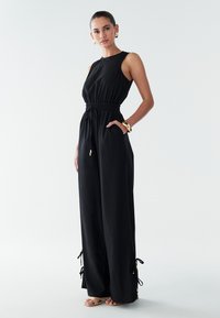 Mono negro con diseño sin mangas, cintura elástica y cordón ajustable. Cuenta con bolsillos laterales y detalles anudados en el tobillo. Textura suave.