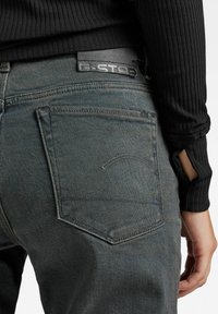 Jeans de mezclilla oscura con un acabado texturizado, que cuentan con un parche de cuero negro de G-Star, costuras en los bolsillos traseros con curva, y una camiseta de manga larga negra acanalada.