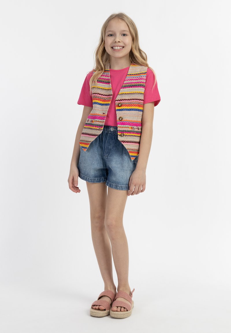 Daugių spalvų liemenė su dryžuotais raštais, derinama su rožine marškinėlių viršuje ir džinsiniais šortais. Modelis vilki rožinius espadrilles.