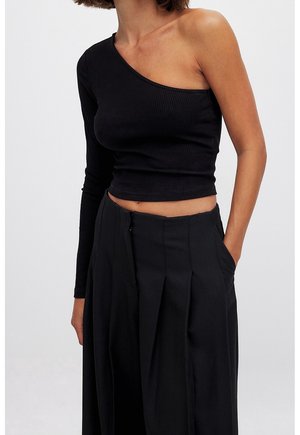 Mujer que lleva un top negro de un solo hombro y manga larga, corto, combinado con pantalones negros plisados de talle alto, con la mano en el bolsillo.