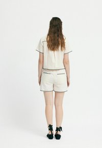 Weißes Baumwollhemd mit schwarzer Einfassung, kombiniert mit weißen Shorts, die kontrastierende Einfassungen aufweisen, und schwarzen Riemchenschuhen. Texturiertes, lässiges Design.