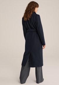 Marinblå ullblandning lång kappa med bälte i midjan, slagkrage och sidoflikar, i kombination med grårandiga byxor.