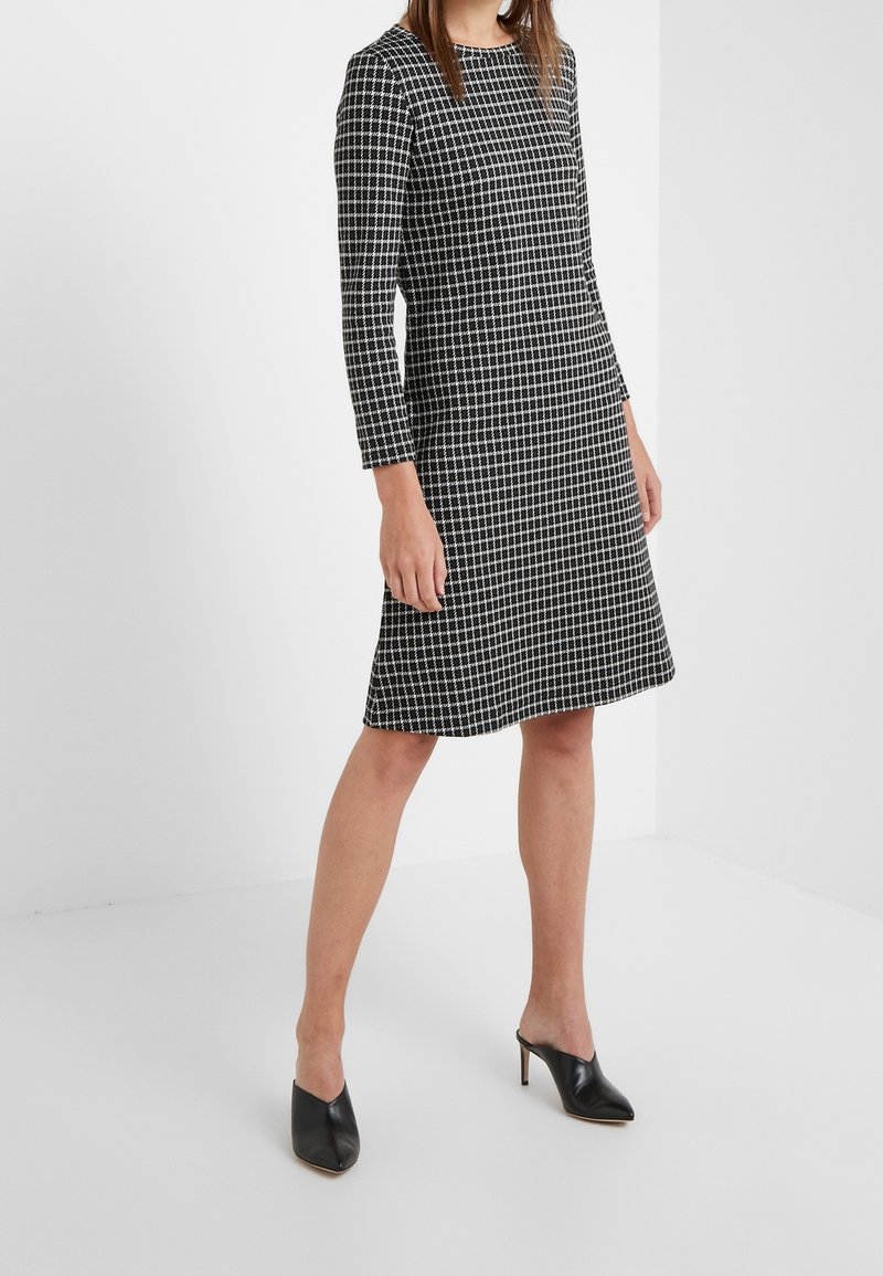 Černobílé kostkované šaty s dlouhými rukávy, kulatým výstřihem, rozšířeným lemem a texturovanou tkaninou. Nosené s černými muly s špičatou špičkou.