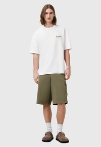 Bijela oversized majica s "ALLSAINTS" logotipom, maslinaste kratke cargo hlače, bijele čarape i smeđe cipele bez vezivanja. Opisan ležeran outfit s prednje strane.