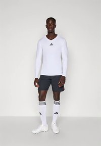 Homem em pé, vestindo uma camisa de mangas largas branca da Adidas, calções pretos, meias brancas até ao joelho da Adidas e sapatilhas brancas, num fundo simples.