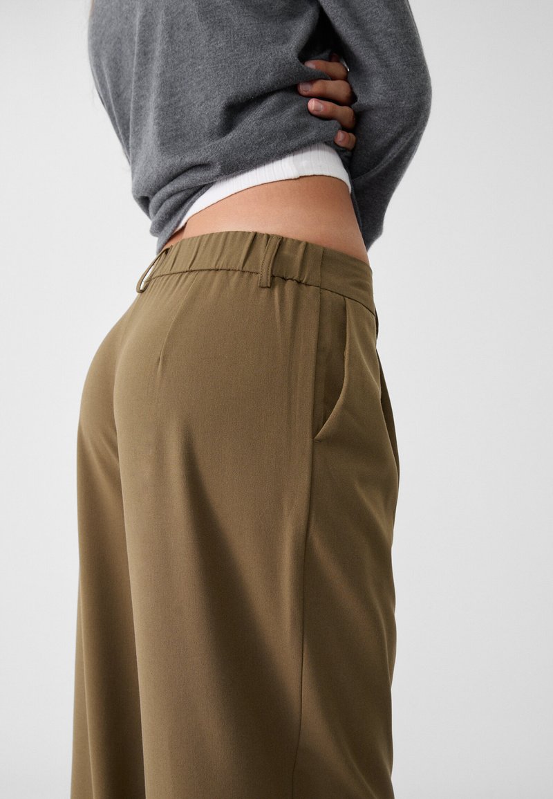 Pantalons vert olive avec une texture lisse, comportant des poches latérales et une ceinture élastique. Associé à un pull en tricot gris.