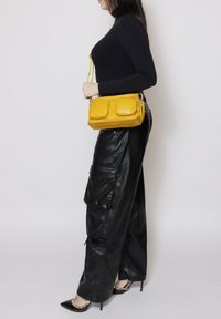 Borsa a tracolla gialla in pelle con due tasche frontali, forma rettangolare e chiusura con cerniera; abbinata a pantaloni cargo neri in pelle e tacchi a punta.