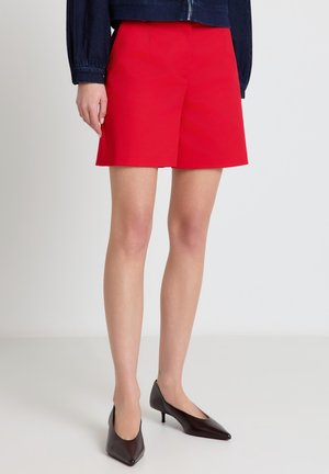 Femme portant un short rouge vif sur mesure, des talons sombres à bout pointu et une veste courte sombre sur un fond uni.