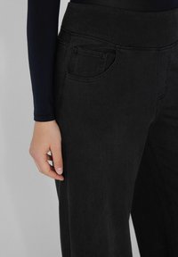 Pantalones de mezclilla negros con una textura suave, cintura de altura media, bolsillo trasero visible y ligero estiramiento. Mano descansando en el costado para escala.