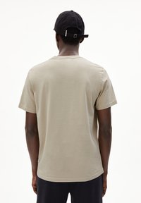 Beige katoenen t-shirt met korte mouwen en een ronde hals, achterstevoren gedragen, in combinatie met een zwarte baseballpet met een verstelbare strap.