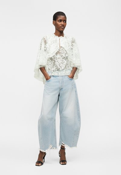 Femme debout, les mains dans les poches, portant un haut en dentelle blanche à motif floral, un jean large bleu clair déchiré et des talons noirs.