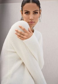 Vrouw met donker haar in een witte, gestructureerde gebreide trui, die haar schouder vasthoudt, met geometrische zilveren oorbellen, op een neutrale achtergrond.