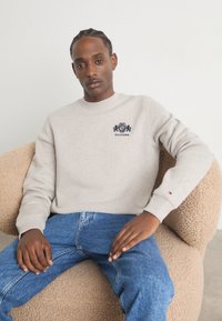 Lys grå sweatshirt med rund hals og broderet marineblå logo. Båret med blå jeans, siddende på en struktureret beige stol.