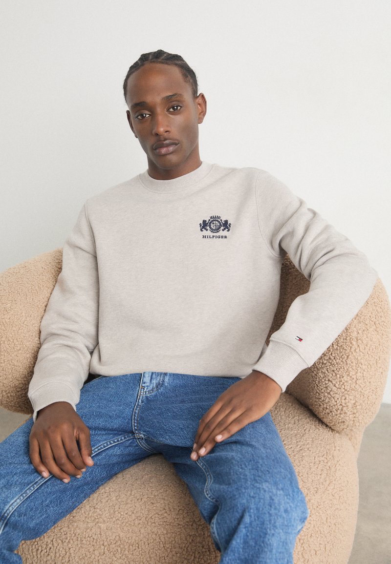 Lys grå sweatshirt med rund hals og broderet marineblå logo. Båret med blå jeans, siddende på en struktureret beige stol.