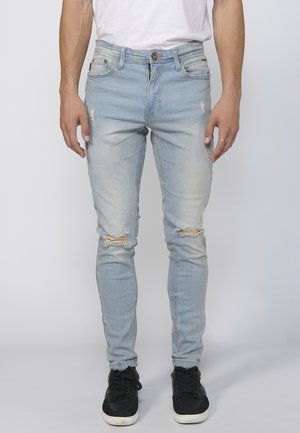 Light blue distressed jeans with rips at the knees, slim fit, five-pocket design, finished with a button and zip fly. Paired with black sneakers.