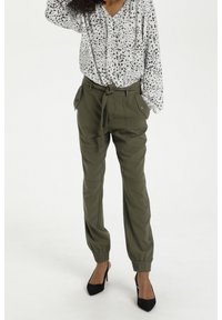 Weißes Hemd mit schwarzem gepunktetem Muster, kombiniert mit olivgrünen, mit einem Gürtel ausgestatteten Hosen mit seitlichen Taschen und elastischen Bündchen, getragen mit schwarzen High Heels.
