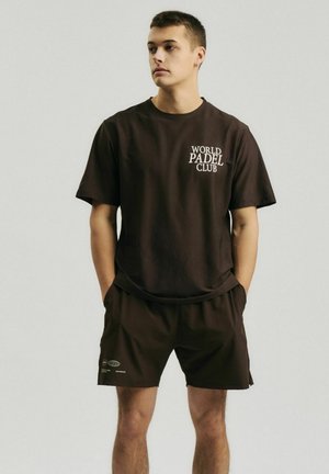 Jeune homme debout, les mains dans les poches, portant un t-shirt marron foncé avec le texte "World Padel Club" et un short assorti sur un fond uni.