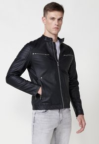 Chaqueta de moto de cuero negro con cuello alzado, bolsillos con cremallera, acentos en los hombros con detalles en relieve y un diseño ajustado.