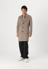 Cappotto in lana beige con revers, tre bottoni, tasche laterali; indossato su una camicia chiara e abbinato a jeans neri e sneakers bianche.
