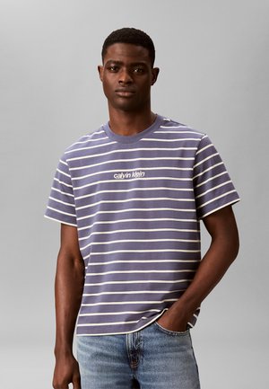 Junger Mann trägt ein kurzärmeliges, marineblau-weiß gestreiftes Calvin Klein T-Shirt und hellblaue Jeans, steht mit einer Hand in der Tasche.