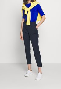 Blå kortärmad topp, gul stickad tröja hängande över axlarna, svarta åtsittande byxor med uppvikt nederkant och vita sneakers.