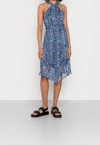 Blå blommig klänning med halterneck, figursydd midja och asymmetrisk fåll. Matchad med kraftiga svarta sandaler med stora spännen.