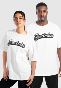 Camisetas blancas de gran tamaño con un logo negro de "Smilodox Athlete", con mangas cortas y un ajuste relajado, combinadas con pantalones deportivos negros.