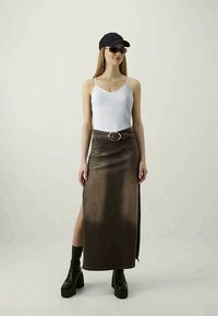 Saia de denim longa em caqui com uma fenda lateral, combinada com uma blusa de alças branca de malha canelada, um boné preto, óculos de sol e botas plataforma grossas pretas.